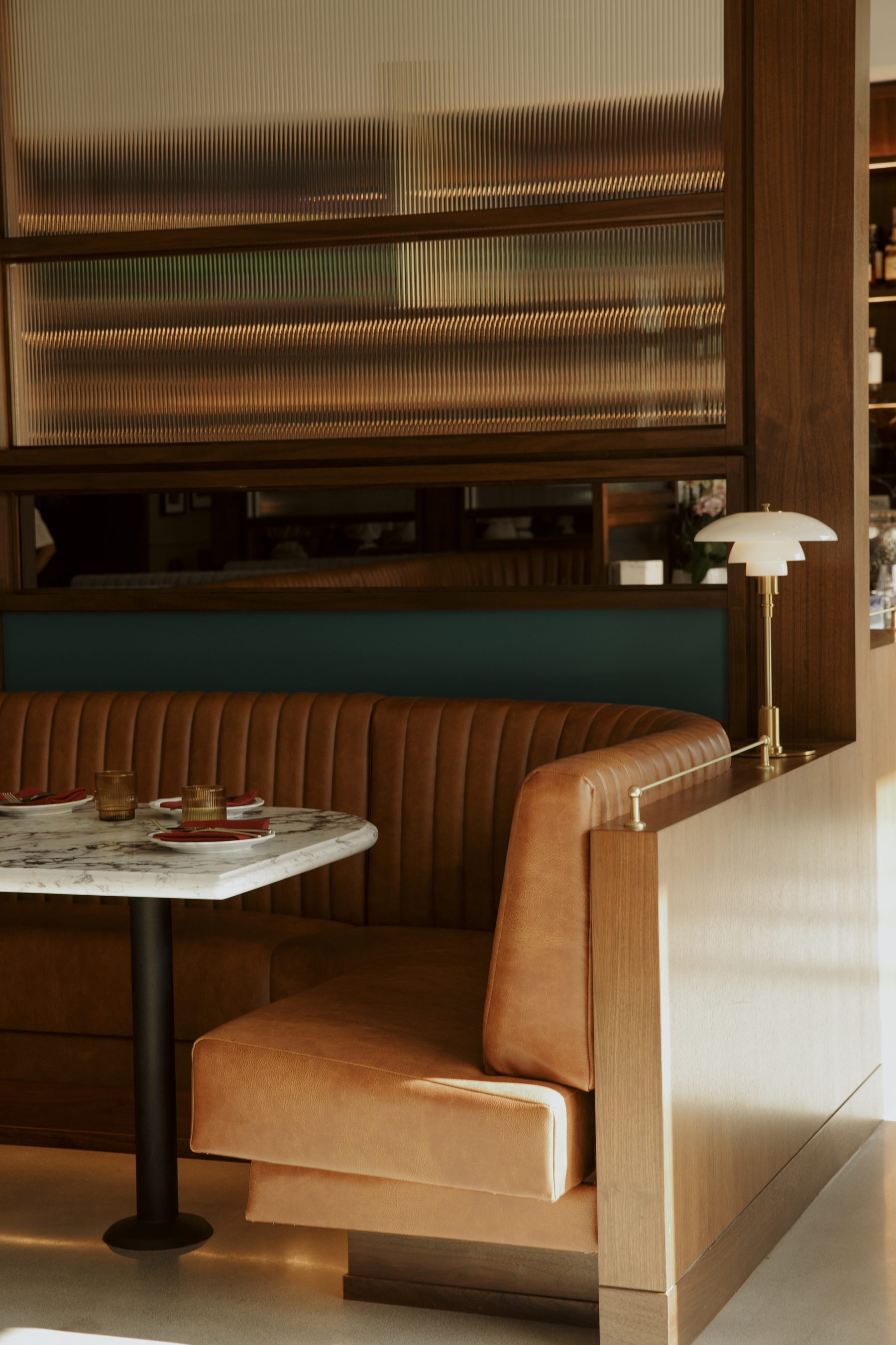 A cognac leather booth at Salina, lit by a brass lamp, with reeded glass and a marble table set with crimson napkins