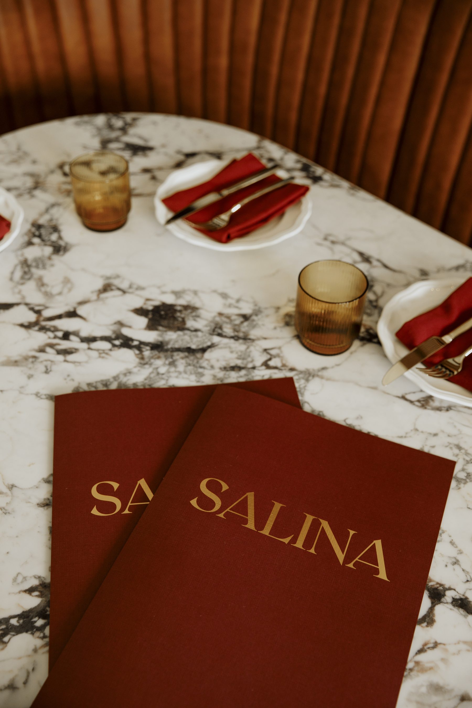 Gold SALINA menus on a white marble table with crimson napkins and amber glassware