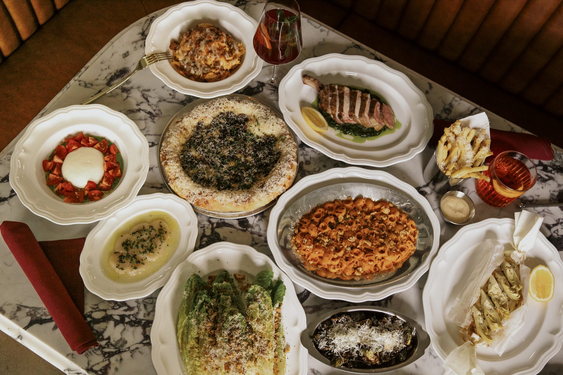 A spread of Salina dishes on marble — lasagna, lamb chop with salsa verde, burrata, vodka rigatoni, the Caesar, and a wood-fired pizza